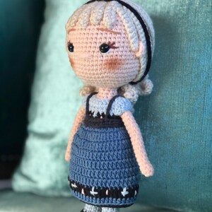 K&ouml;nnte beinhalten: Eine geh&auml;kelte Puppe mit blonden Haaren, einem schwarzen Stirnband und einem blauen Kleid mit schwarzem und wei&szlig;em Besatz. Die Puppe tr&auml;gt schwarze Schuhe.