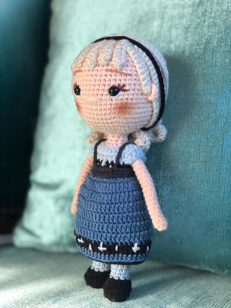 K&ouml;nnte beinhalten: Eine geh&auml;kelte Puppe mit blonden Haaren, einem schwarzen Stirnband und einem blauen Kleid mit schwarzem und wei&szlig;em Besatz. Die Puppe tr&auml;gt schwarze Schuhe.