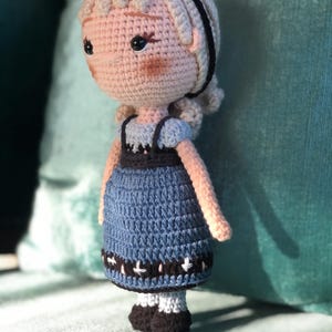 K&ouml;nnte beinhalten: Eine geh&auml;kelte Puppe mit blonden Haaren, die ein blaues Kleid mit schwarzem und wei&szlig;em Besatz tr&auml;gt. Die Puppe hat schwarze Augen und ein rosafarbenes Gesicht.