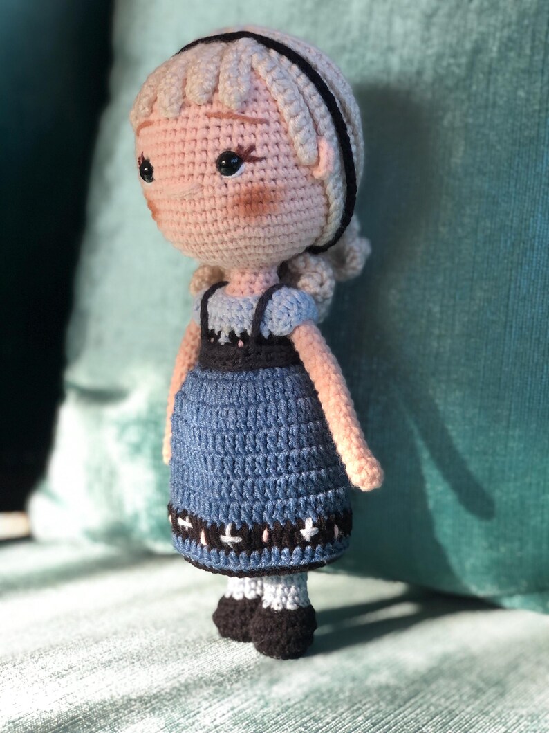 K&ouml;nnte beinhalten: Eine geh&auml;kelte Puppe mit blonden Haaren, die ein blaues Kleid mit schwarzem und wei&szlig;em Besatz tr&auml;gt. Die Puppe hat schwarze Augen und ein rosafarbenes Gesicht.