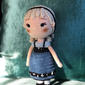 K&ouml;nnte beinhalten: Eine geh&auml;kelte Puppe mit blonden Haaren, einem blauen Kleid mit schwarzem und wei&szlig;em Besatz und schwarzen Schuhen.