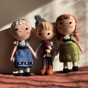 Könnte beinhalten: Drei gehäkelte Puppen, eine mit einem blauen Kleid, eine mit einem grünen Kleid und eine mit einem grauen und braunen Outfit. Die Puppen haben Wollhaare und gestickte Gesichtszüge.