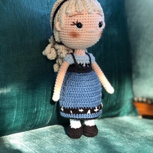 K&ouml;nnte beinhalten: Eine geh&auml;kelte Puppe mit blonden Haaren, einem blauen Kleid und schwarzen Schuhen. Die Puppe sitzt auf einem gr&uuml;nen Stuhl.