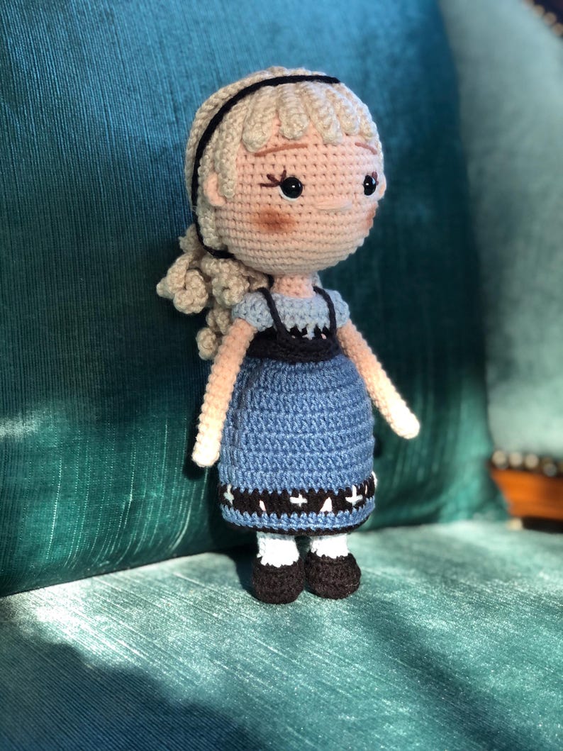 K&ouml;nnte beinhalten: Eine geh&auml;kelte Puppe mit blonden Haaren, einem blauen Kleid und schwarzen Schuhen. Die Puppe sitzt auf einem gr&uuml;nen Stuhl.