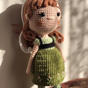 K&ouml;nnte beinhalten: Eine geh&auml;kelte Puppe mit braunen Haaren, einem wei&szlig;en Stirnband, einem gr&uuml;nen Kleid mit schwarzen Akzenten und schwarzen Schuhen. Die Puppe steht auf einer braunen Oberfl&auml;che.