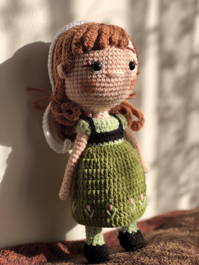 K&ouml;nnte beinhalten: Eine geh&auml;kelte Puppe mit braunen Haaren, einem wei&szlig;en Stirnband, einem gr&uuml;nen Kleid mit schwarzen Akzenten und schwarzen Schuhen. Die Puppe steht auf einer braunen Oberfl&auml;che.
