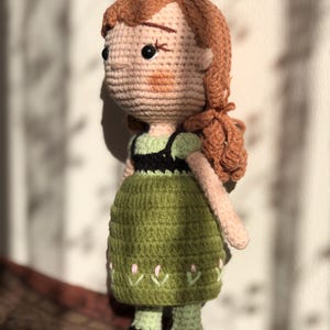 May include: A crocheted doll with brown hair, a green dress, and black shoes. The doll has a pink and green embroidered design on the skirt.