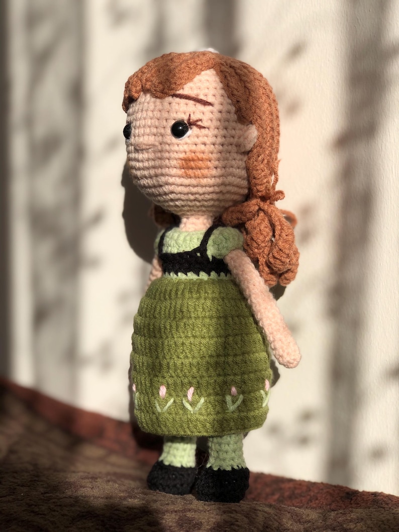 May include: A crocheted doll with brown hair, a green dress, and black shoes. The doll has a pink and green embroidered design on the skirt.