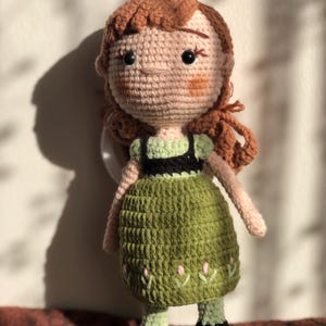 May include: Crocheted doll with brown hair, a green dress, and black shoes. The doll has a smiling face with brown eyes.