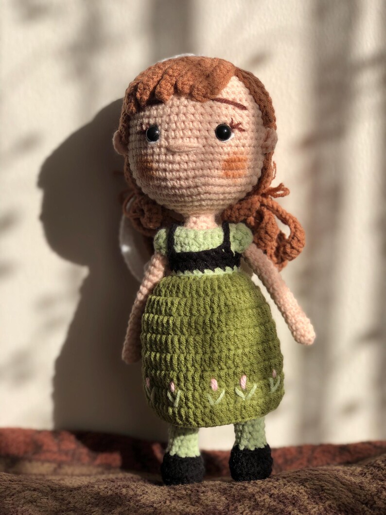 May include: Crocheted doll with brown hair, a green dress, and black shoes. The doll has a smiling face with brown eyes.