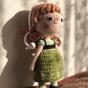May include: A crocheted doll with brown hair, a white headband, and a green dress. The doll is standing on a brown surface.