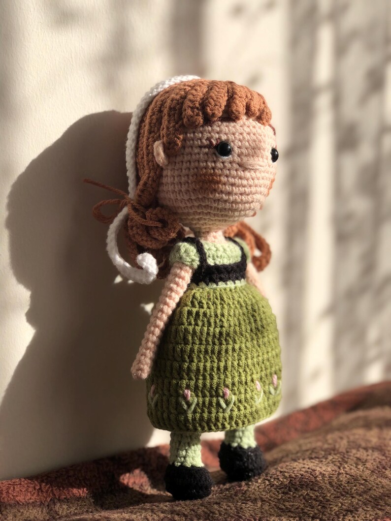 May include: A crocheted doll with brown hair, a white headband, and a green dress. The doll is standing on a brown surface.