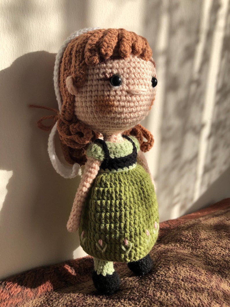 May include: Crocheted doll with brown hair, a white headband, and a green dress with floral embroidery. The doll has black eyes and a pink nose.