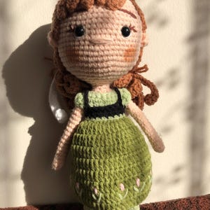 May include: A crocheted doll with brown hair, a green dress, and black shoes. The doll has a pink face with embroidered details.