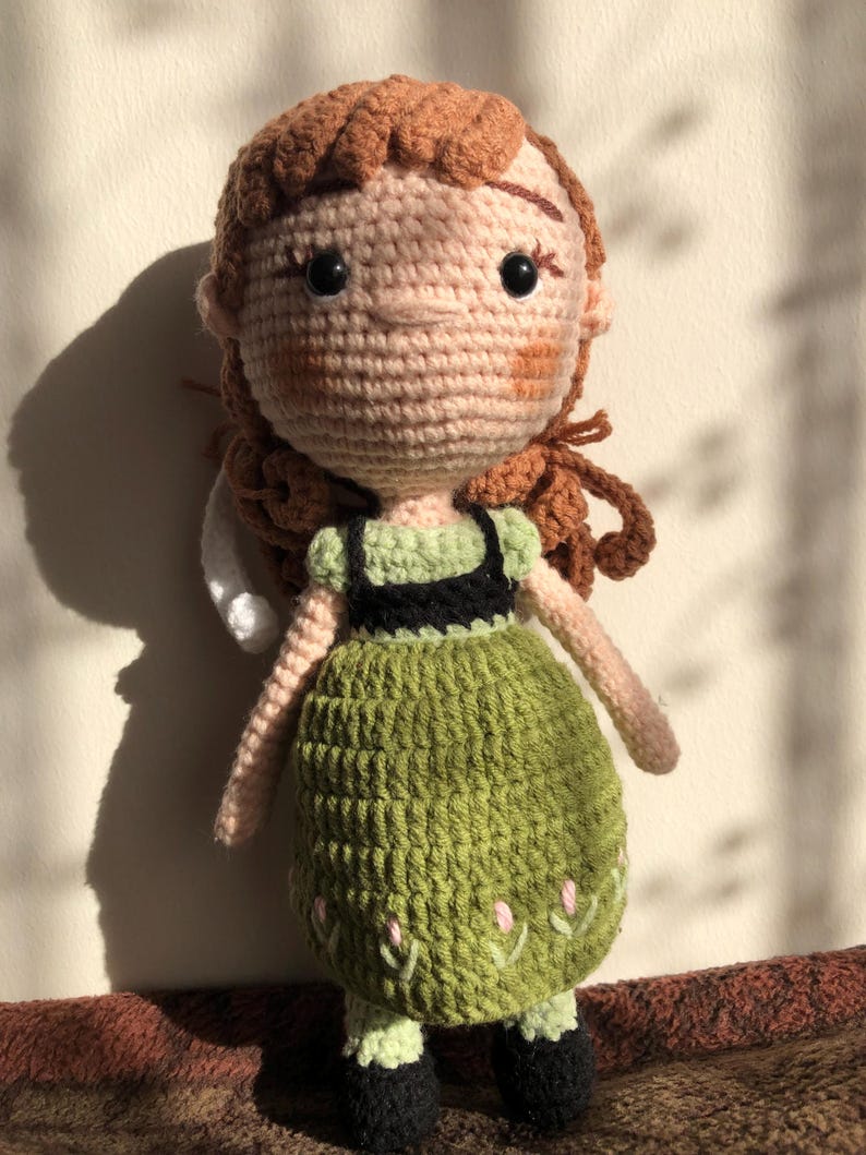 May include: A crocheted doll with brown hair, a green dress, and black shoes. The doll has a pink face with embroidered details.