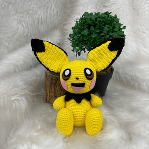 May include: A yellow crocheted Pikachu plush toy with black ears and a black collar. The toy is sitting on a white fluffy surface with a small green potted plant behind it.