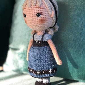 K&ouml;nnte beinhalten: Eine geh&auml;kelte Puppe mit blonden Haaren, einem schwarzen Stirnband und einem blauen Kleid mit wei&szlig;en Akzenten. Die Puppe steht auf einer gr&uuml;nen Oberfl&auml;che.