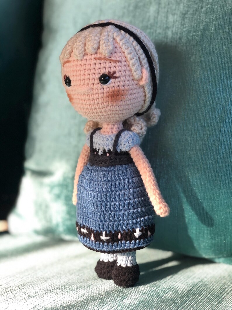 K&ouml;nnte beinhalten: Eine geh&auml;kelte Puppe mit blonden Haaren, einem schwarzen Stirnband und einem blauen Kleid mit wei&szlig;en Akzenten. Die Puppe steht auf einer gr&uuml;nen Oberfl&auml;che.