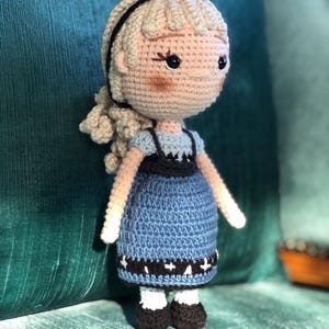 K&ouml;nnte beinhalten: Eine geh&auml;kelte Puppe mit blonden Haaren, einem schwarzen Stirnband und einem blauen Kleid mit wei&szlig;en Akzenten. Die Puppe steht auf einer gr&uuml;nen Oberfl&auml;che.