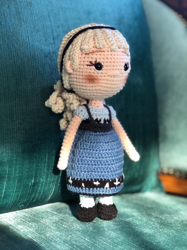 K&ouml;nnte beinhalten: Eine geh&auml;kelte Puppe mit blonden Haaren, einem schwarzen Stirnband und einem blauen Kleid mit wei&szlig;en Akzenten. Die Puppe steht auf einer gr&uuml;nen Oberfl&auml;che.