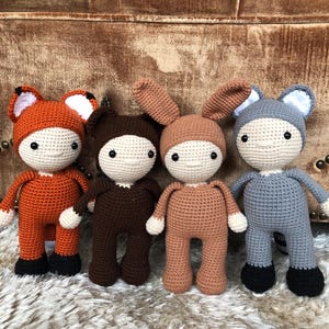 May include: Four crocheted stuffed toys wearing different animal costumes. The animals are a fox, a bear, a bunny, and a raccoon. The toys are all standing on a fluffy rug.