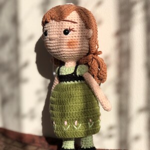 May include: A crocheted doll with brown hair, a green dress, and black shoes. The doll is standing on a brown surface.