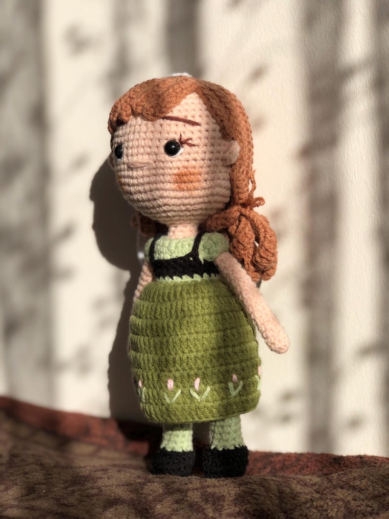 May include: A crocheted doll with brown hair, a green dress, and black shoes. The doll is standing on a brown surface.