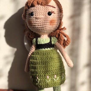 May include: A crocheted doll with brown hair, a green dress, and black shoes. The doll has a light pink face with embroidered details.