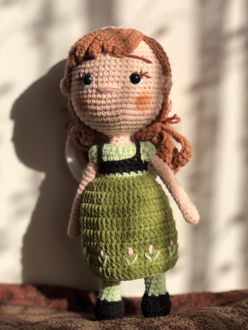 May include: A crocheted doll with brown hair, a green dress, and black shoes. The doll has a light pink face with embroidered details.