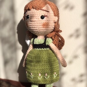 May include: A crocheted doll with brown hair, a green dress with a black bodice, and black shoes. The doll has a stitched-on face with brown eyes and a pink blush.