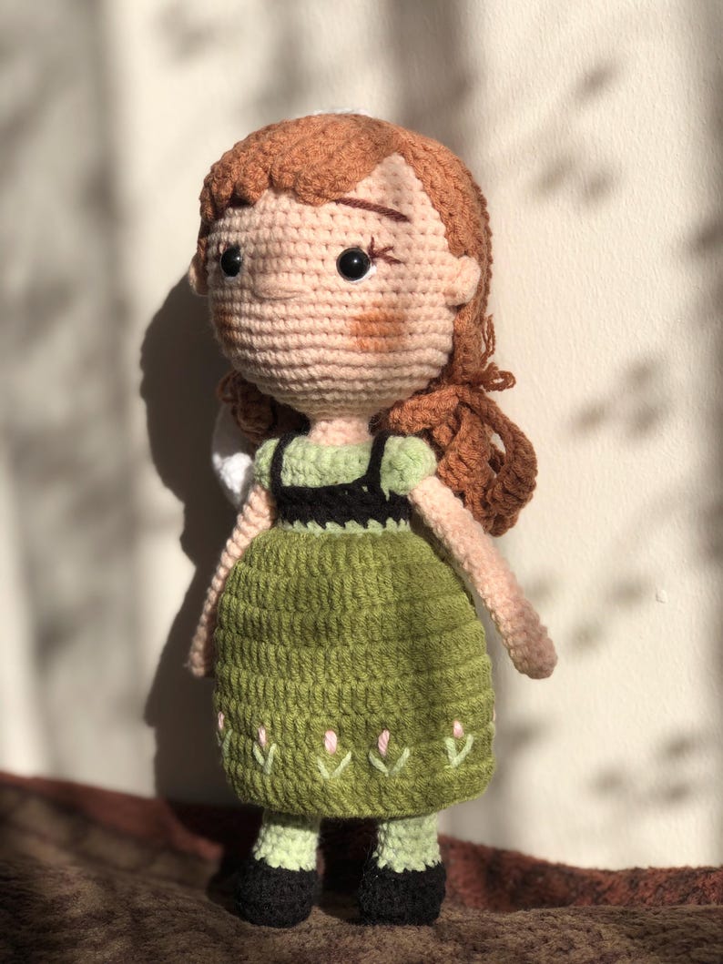 May include: A crocheted doll with brown hair, a green dress with a black bodice, and black shoes. The doll has a stitched-on face with brown eyes and a pink blush.