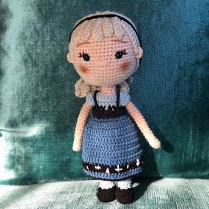 K&ouml;nnte beinhalten: Eine geh&auml;kelte Puppe mit blonden Haaren, einem blauen Kleid mit schwarzem und wei&szlig;em Besatz und schwarzen Schuhen. Die Puppe steht auf einer gr&uuml;nen Oberfl&auml;che.