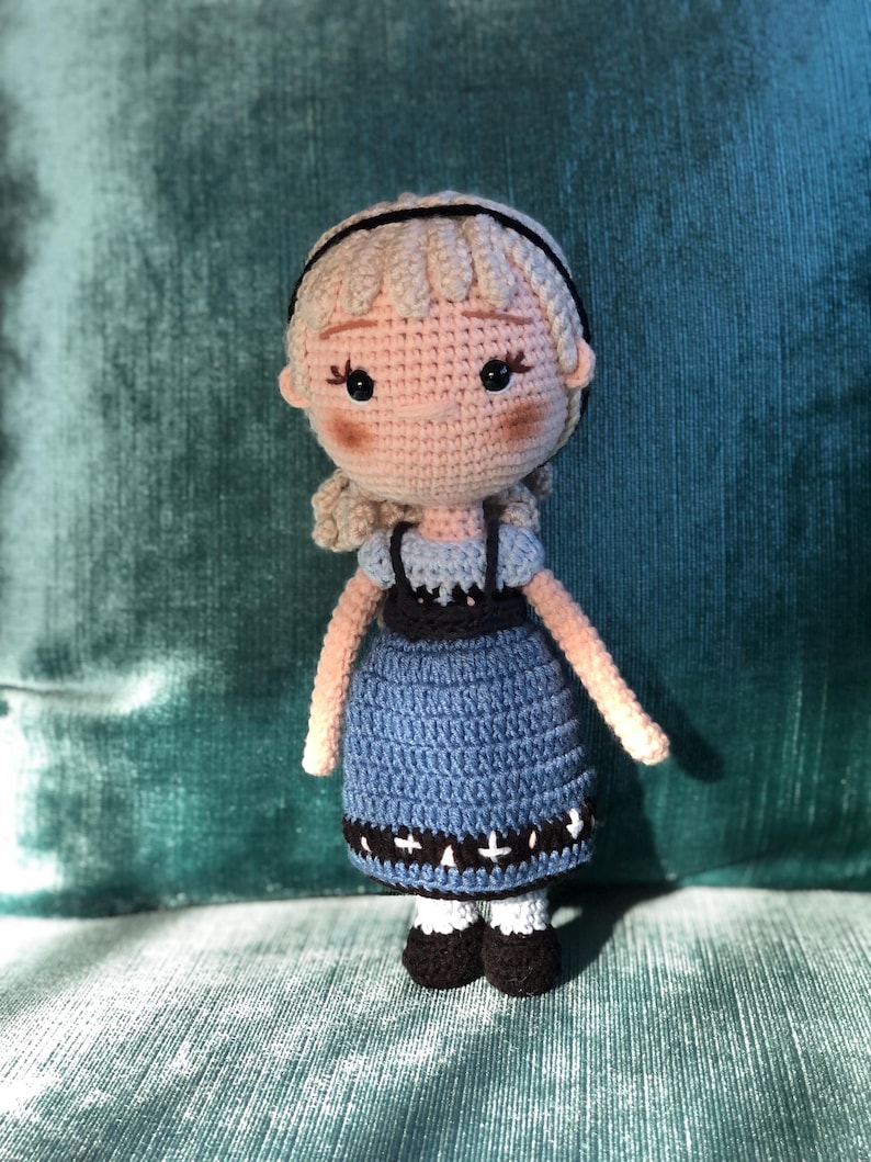 K&ouml;nnte beinhalten: Eine geh&auml;kelte Puppe mit blonden Haaren, einem blauen Kleid mit schwarzem und wei&szlig;em Besatz und schwarzen Schuhen. Die Puppe steht auf einer gr&uuml;nen Oberfl&auml;che.