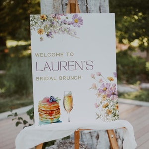 May include: A white sign with a floral design and the text "Welcome to Lauren's Bridal Brunch". The sign is on a wooden easel with a white cloth draped over it.
