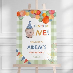 Pode incluir: Uma placa de aniversário em um cavalete com o texto "FUN TO BE ONE!" e "Welcome to Aiden's First Birthday". A placa apresenta uma foto de um bebê usando um chapéu de festa, balões coloridos e um fundo xadrez verde.