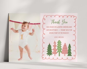 Puede incluir: Una tarjeta fotográfica con un bebé con un body blanco con el número 1 dorado. La tarjeta también presenta un borde rosa con las palabras "Thank You" y un mensaje sobre un país de las maravillas invernal, junto con árboles ilustrados.