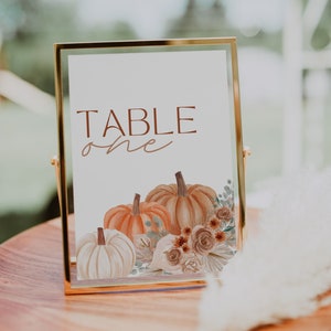 May include: A gold-framed table number sign with the text "TABLE one" and a watercolour illustration of pumpkins and flowers.