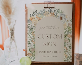 Op de afbeelding: Een aangepast bord met een bloemenontwerp, vastgehouden door een metalen clip op een houten bord. Het bord leest "CUSTOM SIGN" met ruimte voor gepersonaliseerde tekst. Het bord wordt weergegeven op een houten standaard.