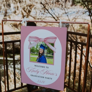 Puede incluir: Un letrero rosa para una fiesta de graduaci&oacute;n con una foto de una graduada con toga y birrete azules. El letrero dice "Welcome to Karly Marie's Graduation Party 7.8.21" y est&aacute; decorado con un lazo de acuarela.