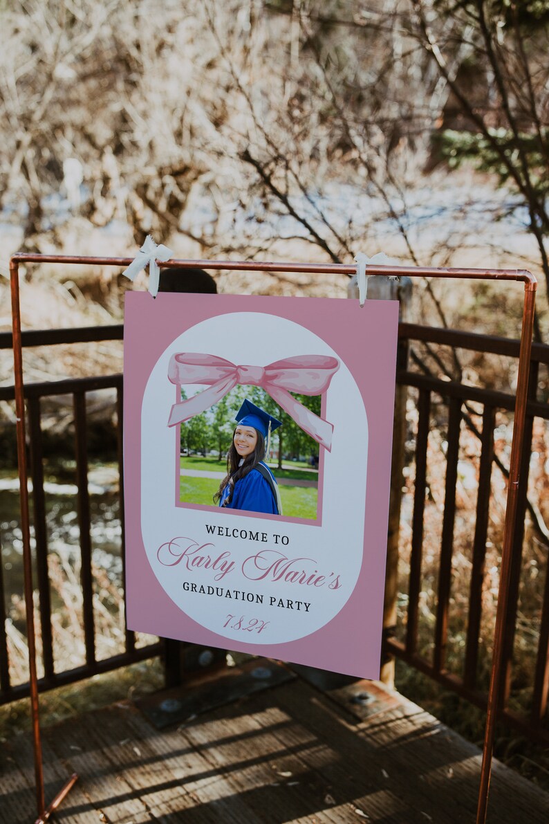 Puede incluir: Un letrero rosa para una fiesta de graduaci&oacute;n con una foto de una graduada con toga y birrete azules. El letrero dice "Welcome to Karly Marie's Graduation Party 7.8.21" y est&aacute; decorado con un lazo de acuarela.