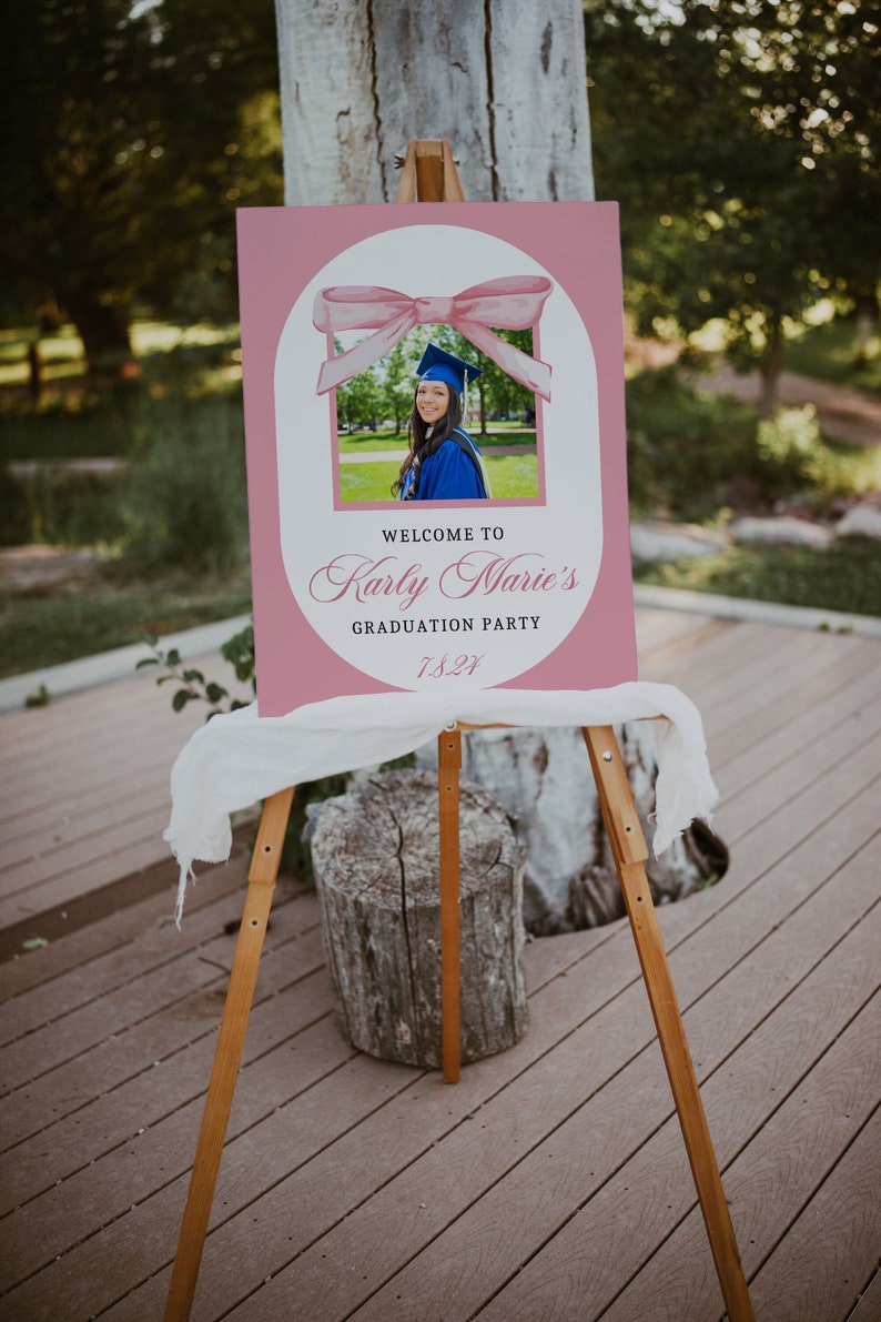 Puede incluir: Un letrero rosa para una fiesta de graduaci&oacute;n sobre un caballete de madera. El letrero presenta una foto de una graduada con toga y birrete azules, con el texto "Welcome to Karly Marie's Graduation Party 7.8.21". Una tela blanca cubre el caballete.