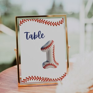 May include: A gold-framed table number sign with a white background and red and white baseball stitching. The number one is a baseball with red stitching. The sign reads "Table 1".
