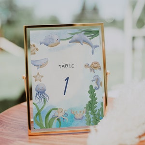 May include: A gold-framed table number card with a blue and white watercolor illustration of sea creatures, including a dolphin, a whale, a starfish, a jellyfish, an octopus, and a crab. The card reads "TABLE 1".