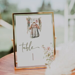 May include: A gold-framed table number card with a watercolor illustration of a dress hanging on a clothesline with flowers. The card says "table 1" in a cursive font.