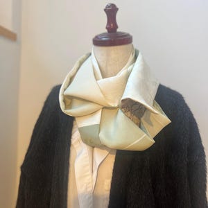 以下が含まれることがあります： クリーム、グリーン、ブラウンの色合いの折り畳まれたスカーフがマネキンの首にかけられています。 スカーフはシルクのような素材でできており、重ねて質感のある効果を生み出しています。 マネキンは白い襟付きシャツの上に黒いカーディガンを着ています。