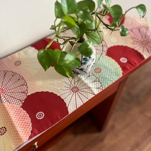 May include: A red table runner with a floral pattern featuring large, white, and pink circles with a gold and red checkered pattern. A green plant in a white and blue ceramic vase is on top of the runner.