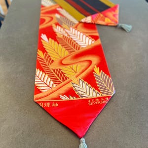 以下が含まれることがあります： 伝統的な日本のデザインが施された赤と金のテーブルランナー。ランナーはリバーシブルで、両端にタッセルが付いています。ランナーのテキストには「Japanese Table Runner」と「Reversible」とあります。