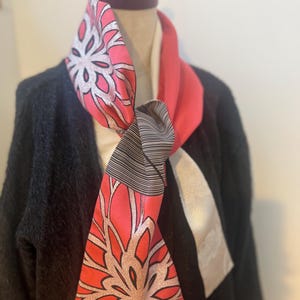 以下が含まれることがあります： ピンク、白、シルバーの色合いの花柄が特徴的な鮮やかなシルクのスカーフ。 スカーフは黒いセーターの上にドレープされており、そのデザインが際立っています。 スカーフには、白と黒のストライプのセクションもあります。