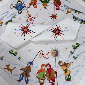 Puede incluir: Servilletas de lino blanco con diseños de explosiones bordadas. Las servilletas están colocadas sobre un mantel blanco con una escena invernal bordada con figuras, árboles y estrellas. El bordado presenta colores rojo, azul, verde y dorado.