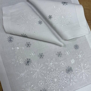 May include: White linen table runner and napkins set, embroidered with snowflake designs in white and grey. The napkins are folded, displaying the snowflake embroidery. The runner and napkins are on a wooden surface.